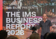 IMS Business Report: Die elektronische Musik hat sich von Corona erholt Der Analyst Mark Mulligan steht etwas seitlich auf einer Bühne und präsentiert den IMS Business Report 2023. Hinter ihm an der Wand ist das Titelbild dieses Berichts an die Wand projiziert. Es handelt sich um eine Hafenaufnahme der Stadt Ibiza bei Nacht.